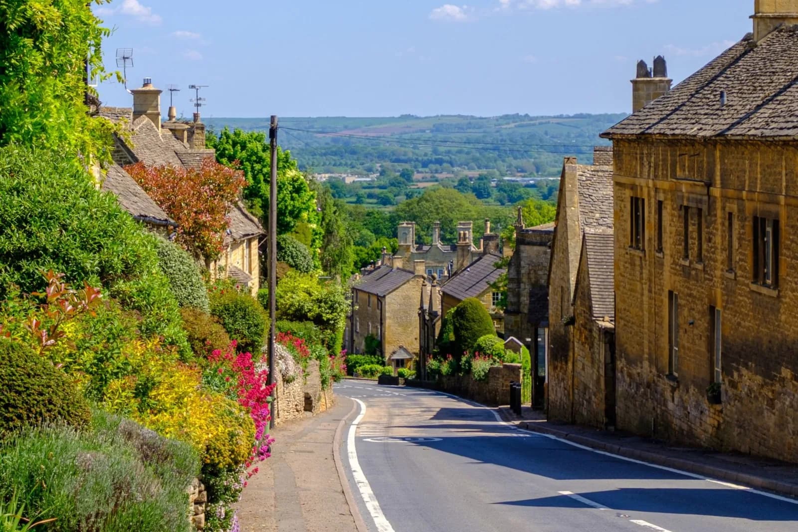 Rue en pente bordée de maisons en pierre dorée et de massifs fleuris, dans un village des Cotswolds