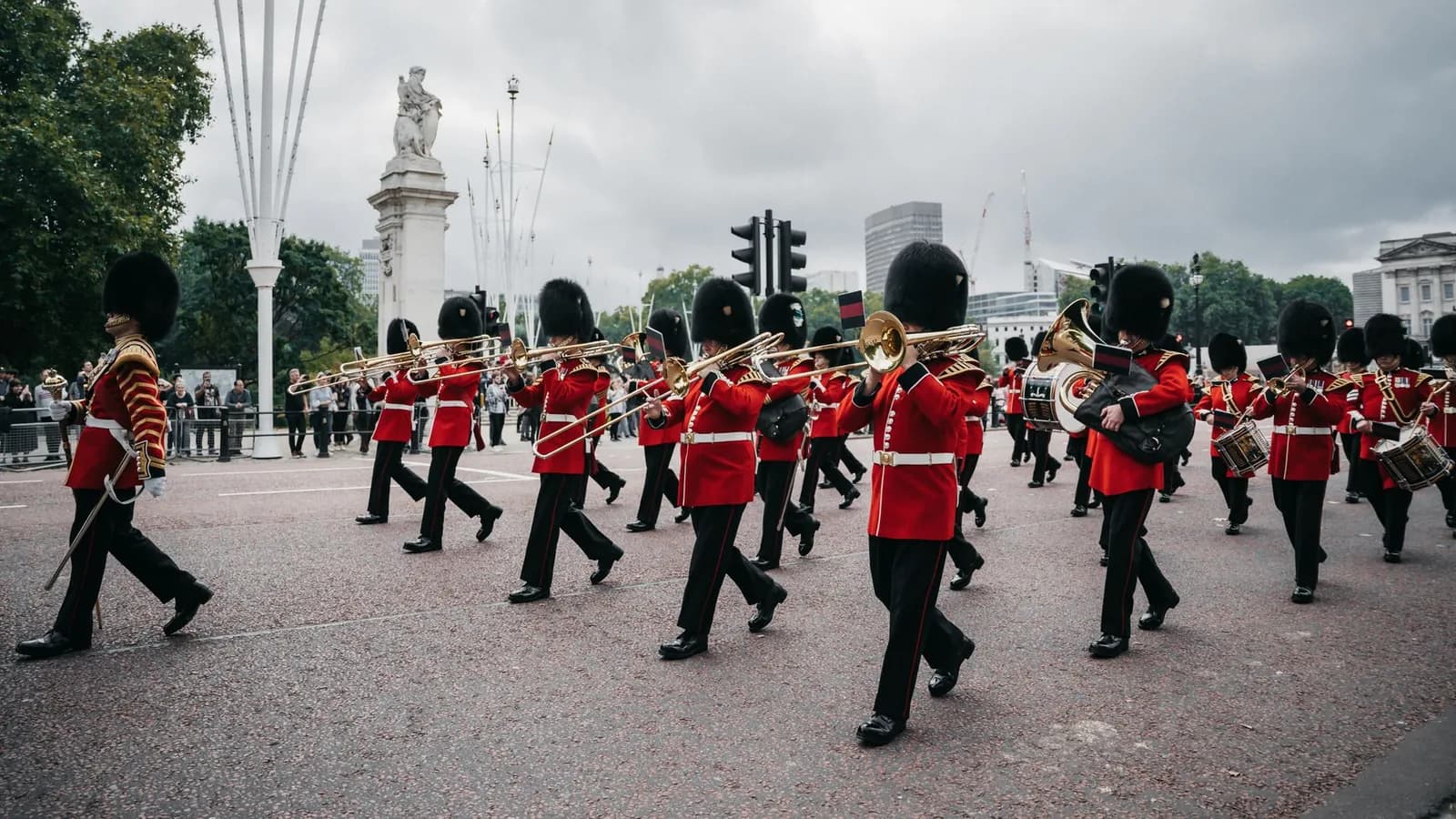 Fanfare de gardes en tunique rouge et bonnet à poil défilant sur une large chaussée rougeâtre, avec un monument de pierre blanche à l'arrière-plan