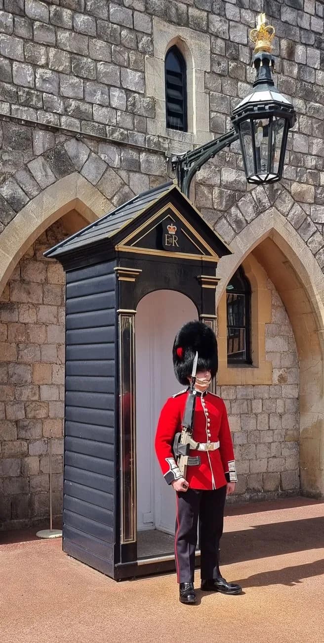 Garde en tunique rouge et bonnet à poil devant sa guérite noire, contre un mur de pierre à arcades