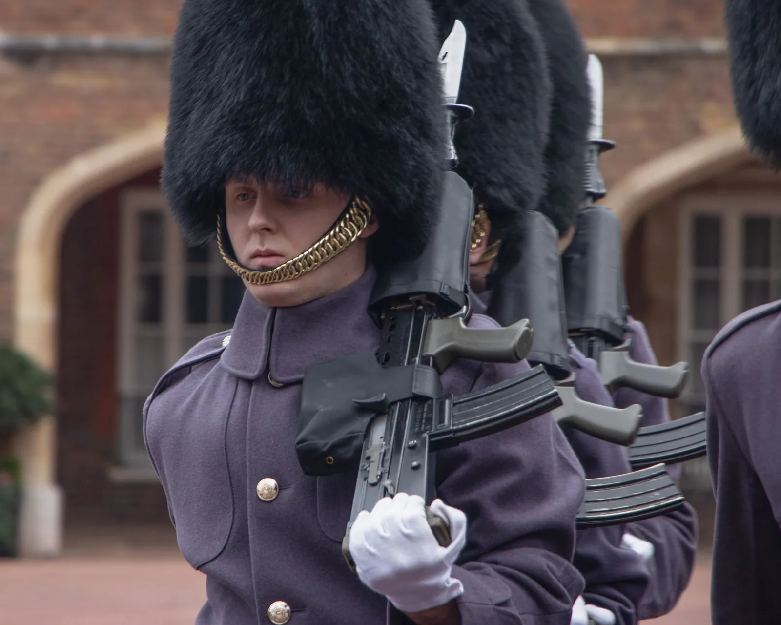 Gardes en capote grise, bonnet à poil et fusil à l'épaule, marchant en formation devant des arcades de brique