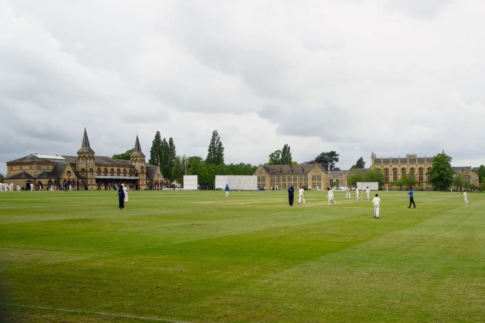 Match de cricket sur une grande pelouse tondue, joueurs en blanc devant des bâtiments en pierre