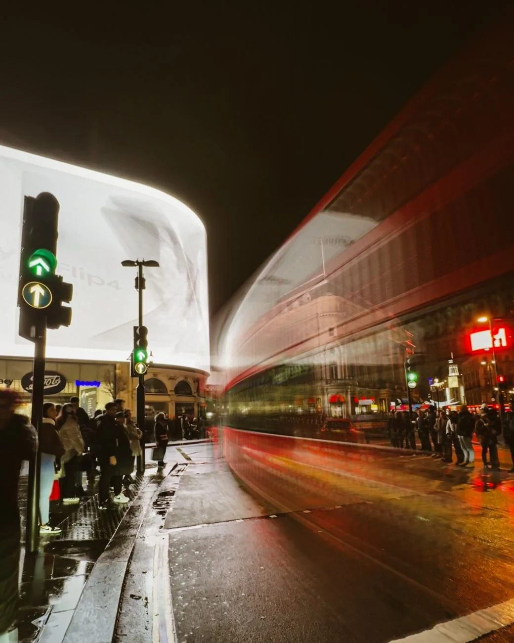 Piccadilly Circus de nuit sous la pluie, un bus rouge en mouvement devant les écrans