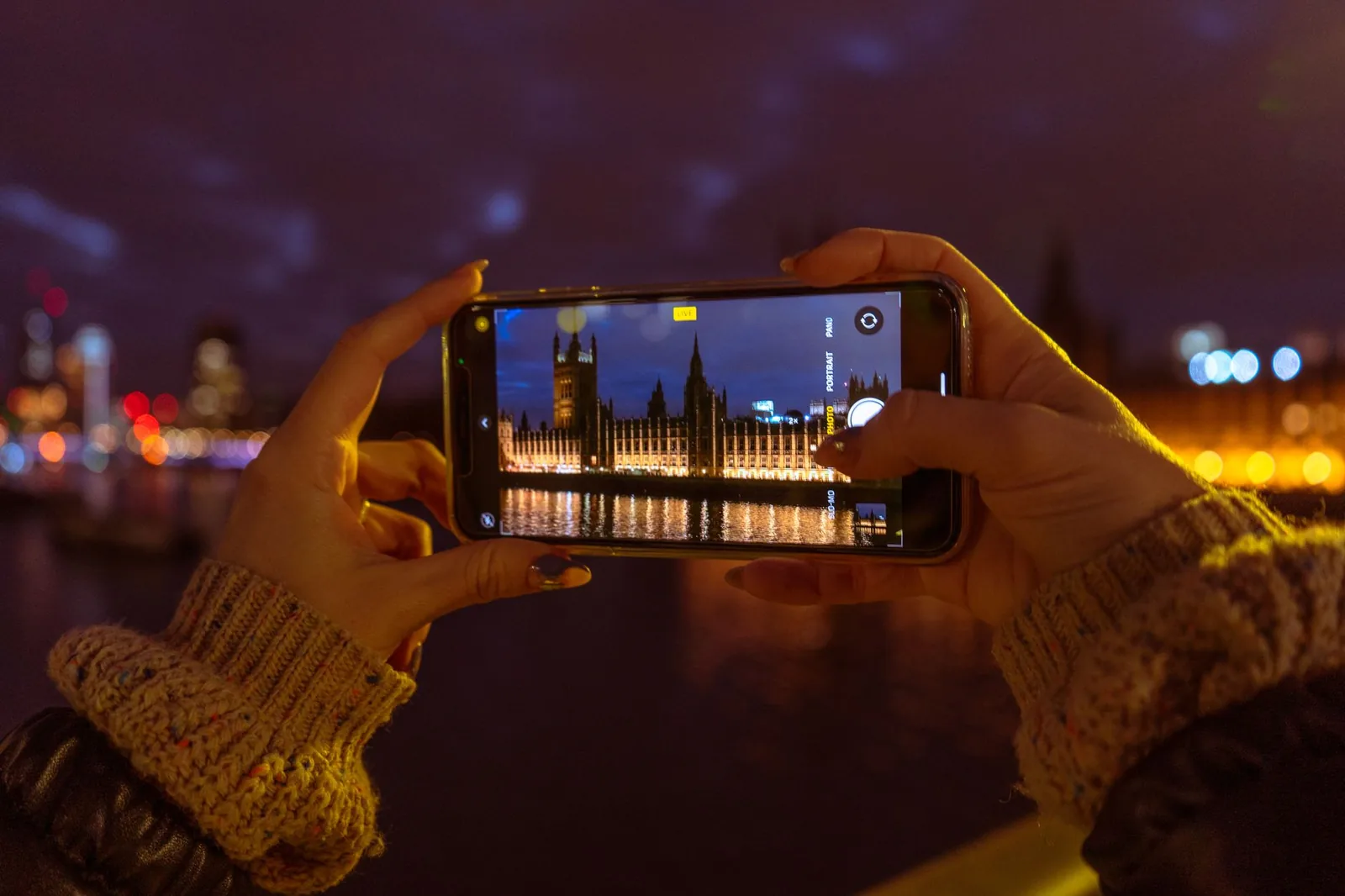 Mains tenant un smartphone qui photographie le palais de Westminster et Big Ben de nuit, au bord de la Tamise à Londres
