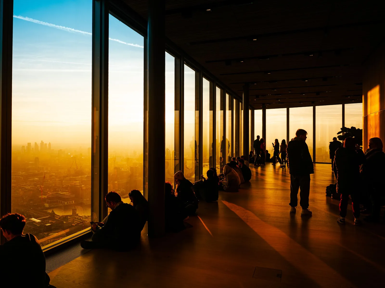 Visiteurs assis et debout devant les grandes baies vitrées de l'observatoire du Shard, au coucher du soleil